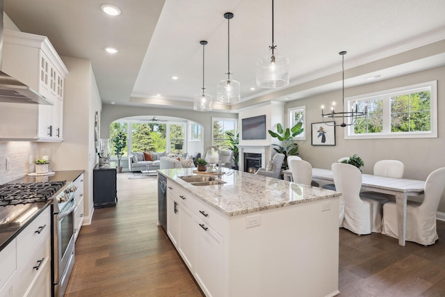 kitchen with a center island with sink, a raised ceiling, stainless steel gas range, wall chimney range hood, and dark wood-type flooring