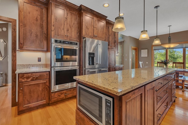 kitchen with hanging light fixtures, appliances with stainless steel finishes, brown cabinetry, a kitchen island, and light stone countertops