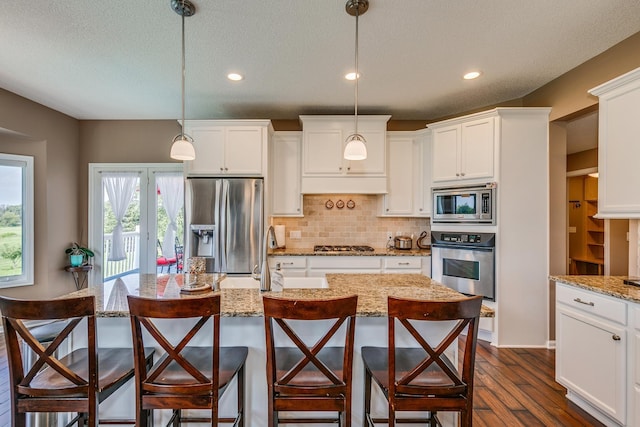 kitchen with dark wood-type flooring, light stone counters, pendant lighting, stainless steel appliances, and a kitchen island with sink