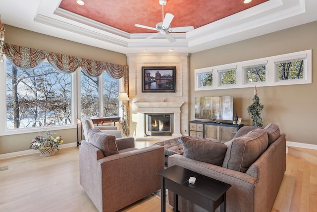 living room featuring a fireplace, ornamental molding, ceiling fan, a tray ceiling, and light wood-type flooring