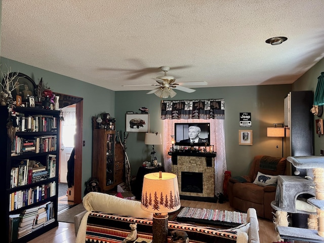 living room featuring wood-type flooring, a stone fireplace, a textured ceiling, and ceiling fan