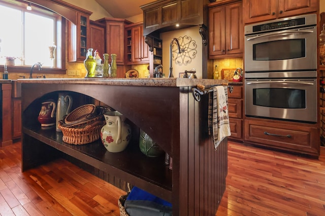 kitchen with glass insert cabinets, stainless steel double oven, dark wood-type flooring, and backsplash