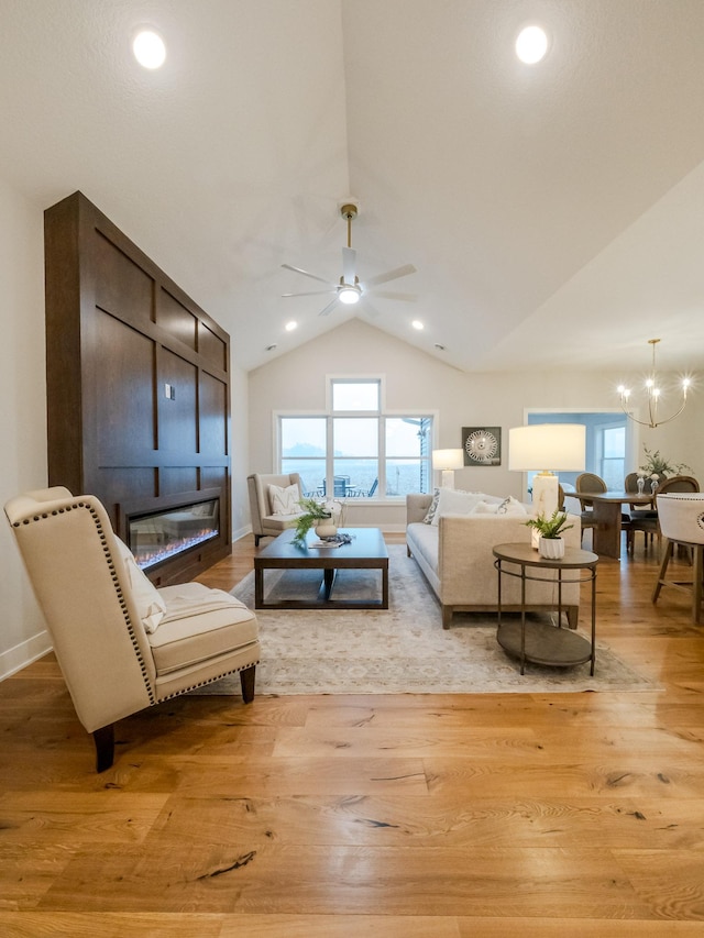 living room with light wood finished floors, ceiling fan with notable chandelier, recessed lighting, a glass covered fireplace, and high vaulted ceiling