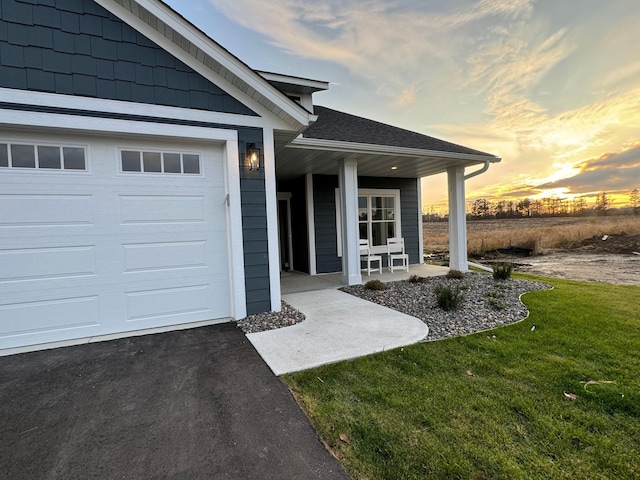 exterior entry at dusk featuring driveway, a porch, an attached garage, a shingled roof, and a lawn