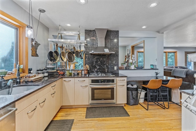 kitchen with wall chimney exhaust hood, open floor plan, stainless steel appliances, light wood-type flooring, and pendant lighting