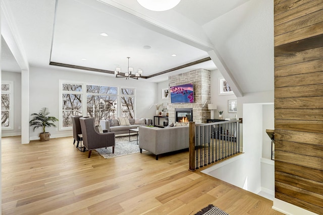 living room with a chandelier, a stone fireplace, a raised ceiling, and light wood-style floors