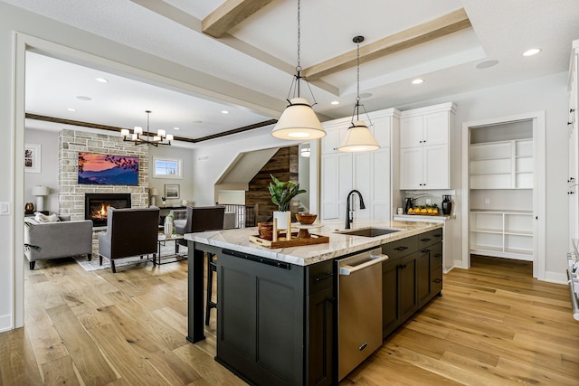 kitchen with light wood finished floors, stainless steel dishwasher, white cabinets, a sink, and a stone fireplace