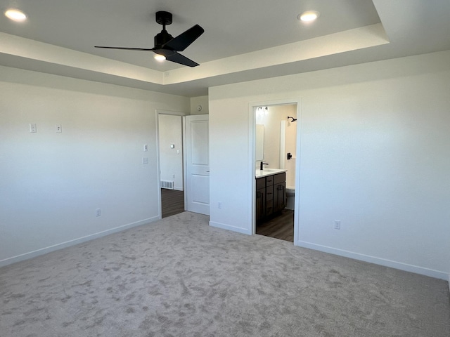 unfurnished bedroom featuring ensuite bath, a tray ceiling, dark colored carpet, and ceiling fan