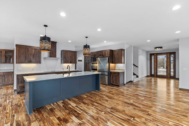 kitchen with wall chimney exhaust hood, hanging light fixtures, hardwood / wood-style floors, an island with sink, and appliances with stainless steel finishes