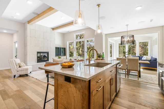 kitchen with a tiled fireplace, beamed ceiling, a kitchen island with sink, light hardwood / wood-style floors, and sink