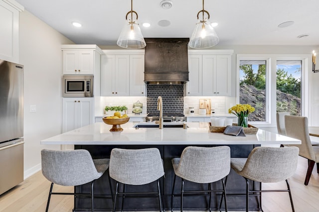 kitchen featuring light hardwood / wood-style flooring, appliances with stainless steel finishes, premium range hood, an island with sink, and decorative backsplash