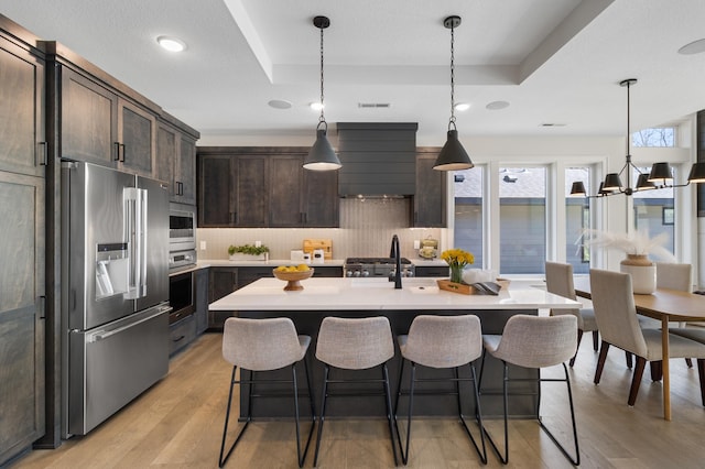 kitchen with a kitchen island with sink, dark brown cabinets, backsplash, appliances with stainless steel finishes, and light hardwood / wood-style floors