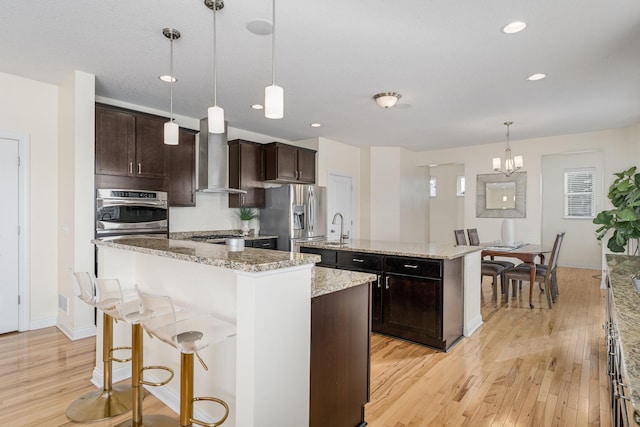 kitchen with wall chimney range hood, an island with sink, light wood-style flooring, and stainless steel fridge with ice dispenser