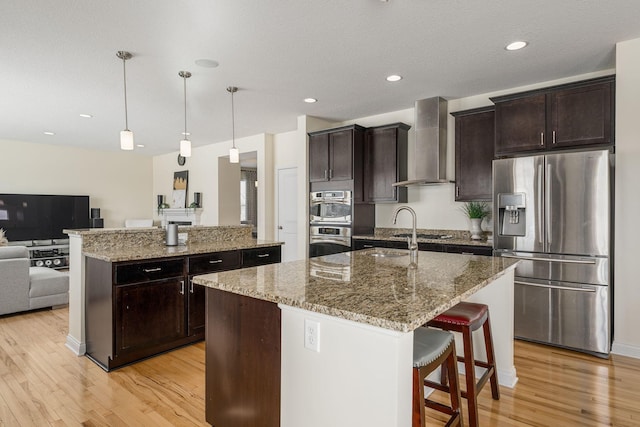 kitchen featuring light wood-style flooring, stainless steel appliances, open floor plan, wall chimney exhaust hood, and a center island with sink