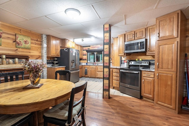 kitchen featuring brown cabinets, light wood-style flooring, a drop ceiling, wooden walls, and appliances with stainless steel finishes