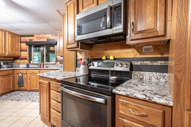 kitchen with brown cabinetry, light stone counters, light tile patterned flooring, stainless steel appliances, and a sink