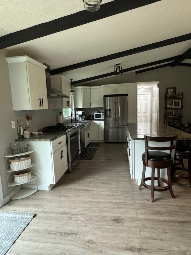 kitchen with dark countertops, lofted ceiling with beams, appliances with stainless steel finishes, white cabinets, and wall chimney range hood