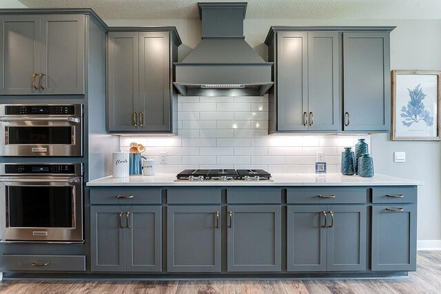 kitchen with backsplash, appliances with stainless steel finishes, wood-type flooring, custom range hood, and gray cabinetry