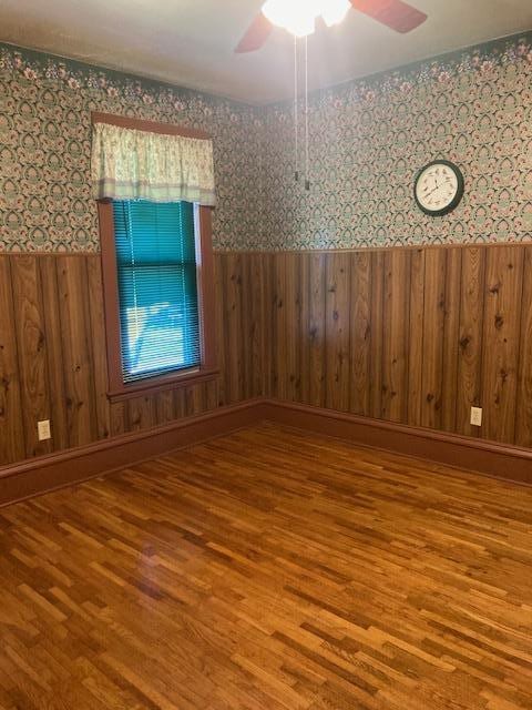 spare room featuring a ceiling fan, wood finished floors, wood walls, wainscoting, and wallpapered walls