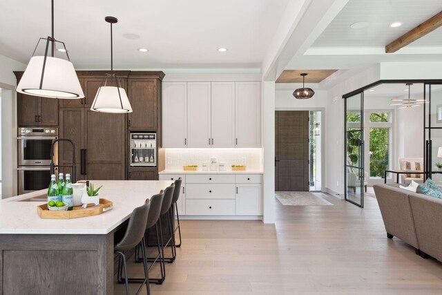 kitchen featuring an island with sink, double oven, beam ceiling, white cabinets, and light hardwood / wood-style floors