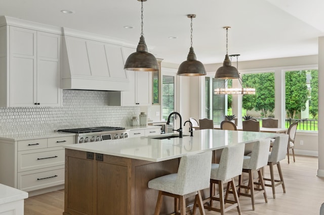 kitchen featuring a large island with sink, white cabinets, sink, decorative light fixtures, and custom range hood