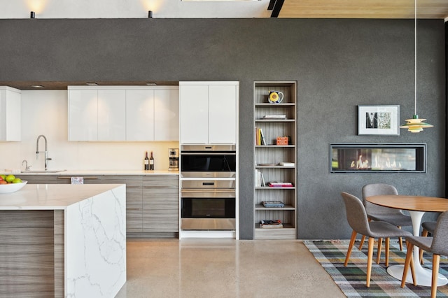 kitchen featuring light stone counters, double oven, sink, white cabinetry, and decorative light fixtures