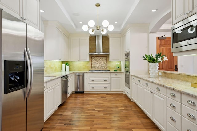 kitchen with appliances with stainless steel finishes, decorative backsplash, hanging light fixtures, a tray ceiling, and wall chimney range hood