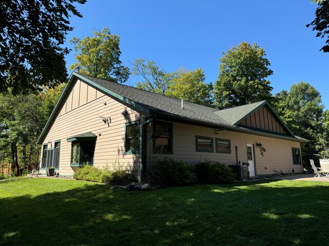 view of front of house with cooling unit, board and batten siding, a front yard, a shingled roof, and a patio area