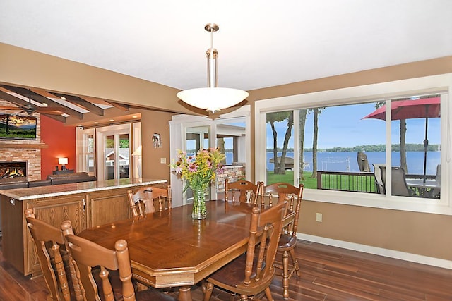 dining area featuring a large fireplace, dark wood-type flooring, a water view, baseboards, and beam ceiling