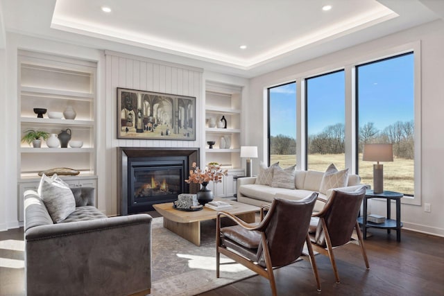 living area featuring built in features, a raised ceiling, dark wood-style flooring, and a glass covered fireplace