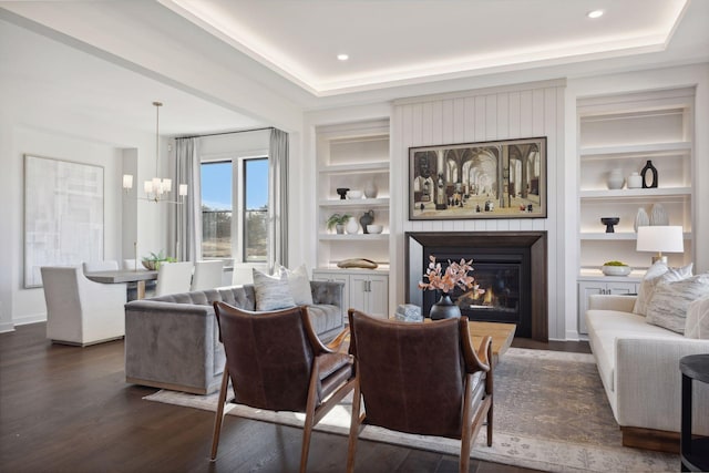 living room with a raised ceiling, a glass covered fireplace, dark wood-type flooring, built in shelves, and a chandelier