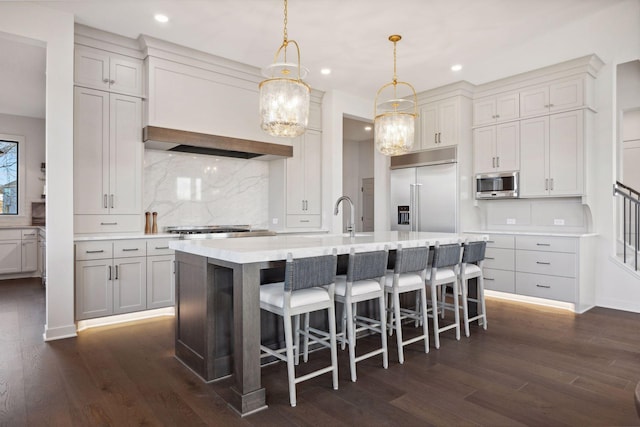 kitchen featuring an island with sink, built in appliances, light countertops, a kitchen bar, and pendant lighting