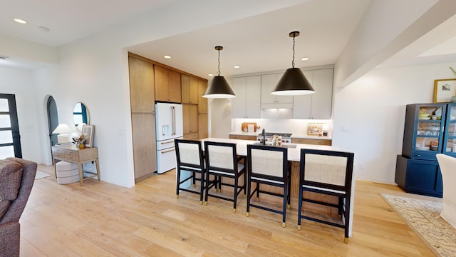 kitchen featuring pendant lighting, light wood-type flooring, white refrigerator with ice dispenser, an island with sink, and white cabinetry