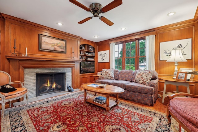 living room featuring built in shelves, a warm lit fireplace, wood finished floors, wooden walls, and ceiling fan