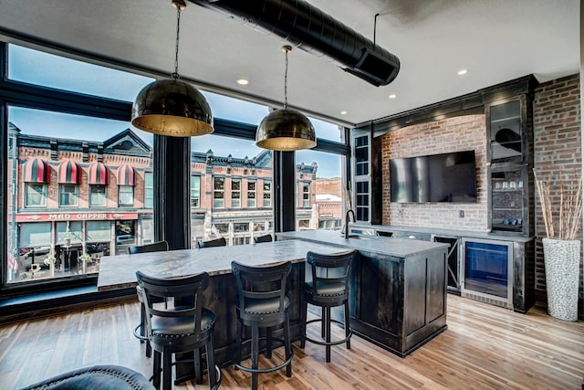 kitchen with wine cooler, an island with sink, pendant lighting, and light wood-type flooring