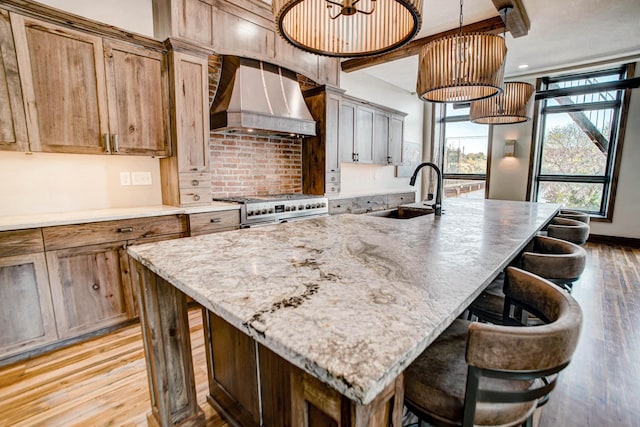 kitchen with light wood-type flooring, stainless steel stove, a kitchen island with sink, and wall chimney range hood