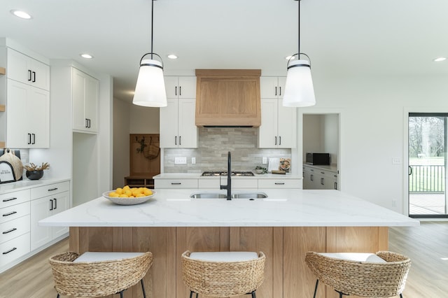 kitchen with light wood-style flooring, premium range hood, a sink, white cabinets, and backsplash