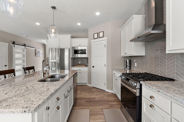 kitchen featuring a sink, a barn door, stainless steel appliances, white cabinets, and wall chimney range hood