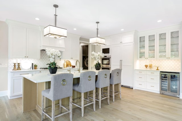 kitchen with light wood-type flooring, white cabinetry, beverage cooler, an island with sink, and hanging light fixtures