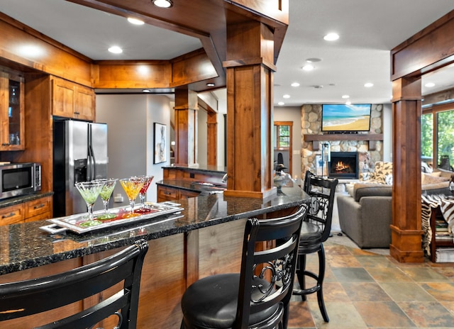 kitchen featuring ornate columns, appliances with stainless steel finishes, a fireplace, dark stone countertops, and a breakfast bar area