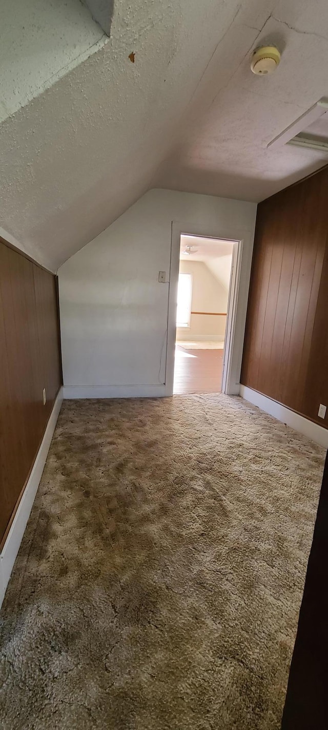 bonus room with wooden walls, a textured ceiling, lofted ceiling, and dark carpet