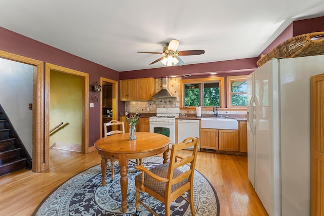 kitchen with white appliances, wall chimney exhaust hood, sink, ceiling fan, and light wood-type flooring