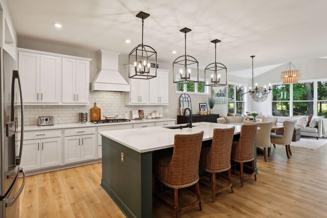 kitchen with custom exhaust hood, hanging light fixtures, sink, a kitchen island with sink, and white cabinetry