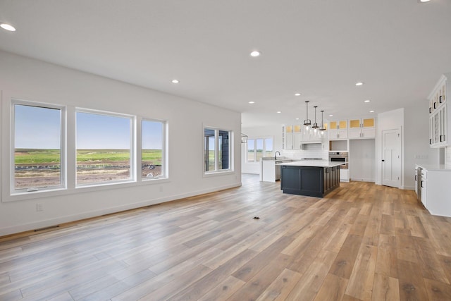 kitchen featuring stainless steel oven, a kitchen island with sink, white cabinets, light hardwood / wood-style floors, and hanging light fixtures