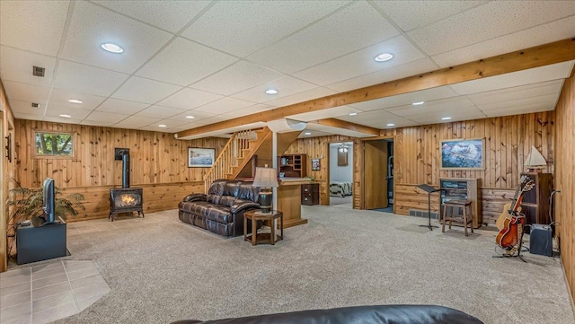 carpeted living room with a drop ceiling, wood walls, and a wood stove