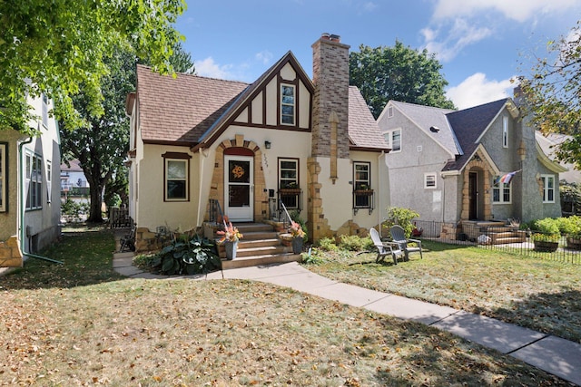 tudor house with stucco siding, roof with shingles, a chimney, and fence