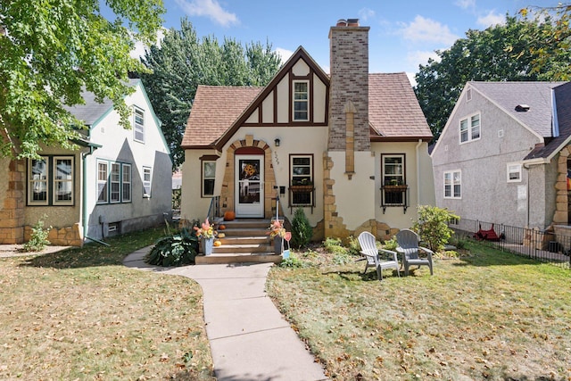 tudor home featuring stucco siding, fence, roof with shingles, and a chimney