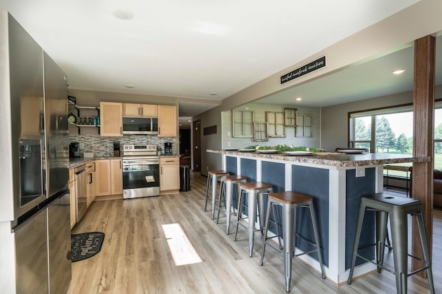 kitchen featuring light brown cabinetry, a breakfast bar area, decorative backsplash, appliances with stainless steel finishes, and open shelves