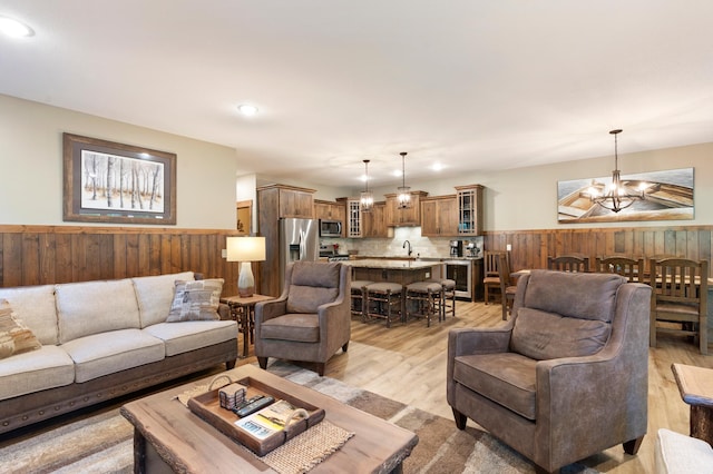 living room with wooden walls, light hardwood / wood-style floors, a notable chandelier, sink, and wine cooler