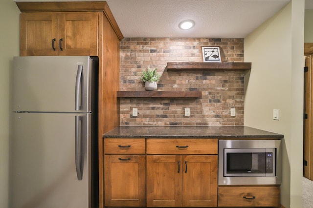 kitchen featuring a textured ceiling, dark stone counters, brown cabinets, stainless steel appliances, and tasteful backsplash